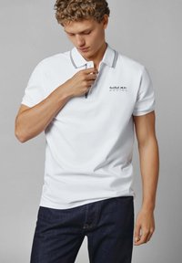 Balta polo marškiniai su juodu apykakle ir plokštele, pristatantys "Red Bull Racing" logotipą kairėje krūtinės pusėje. Nenešiojami su tamsiai mėlynomis džinsais.