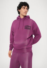 Ljubičasta dukserica s kapuljačom i vezicom, sa crnim logotipom "Aries". Mekani materijal s opuštenim krojem, u kombinaciji s odgovarajućim ljubičastim hlačama.