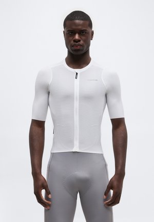 Bărbat purtând un tricou de ciclism alb, cu mânecă scurtă și material plasă, și pantaloni scurți gri căptușiți pentru ciclism, stând pe un fundal neutru.