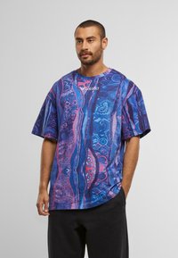 Mand med kort hår og skæg iført en farverig oversized T-shirt i blå og lilla mønster med teksten "COLUCCI", står med den ene hånd i lommen.