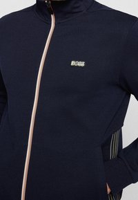 Mörkblå zip-up jacka i mjukt tyg med hög krage, prydd med en broderad "BOSS"-logotyp och färgglada sidstripe.