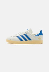 White leather sneakers with blue accents and three stripes, featuring a textured sole, a cushioned collar, and a gold logo on the heel.