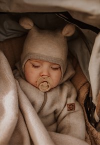 Combineu bej cu fermoar, textură moale și etichetă maro; o căciulă asortată cu urechi de urs. Un suzetă este ținută de un copil adormit.