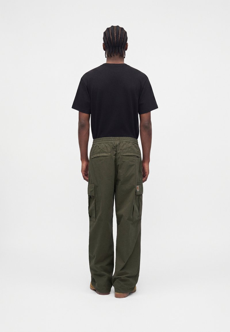 Mann mit geflochtenem Haar, der ein schwarzes T-Shirt und lockere olivgrüne Cargohosen trägt, steht mit dem Rücken zu einem schlichten weißen Hintergrund.