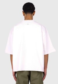 Oversized white cotton t-shirt with short sleeves and a crew neck, features a small label near the back neckline, paired with olive green pants.