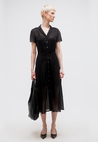 Γυναίκα με κοντά ξανθά μαλλιά που φοράει διάφανο μαύρο μίντι φόρεμα με κουμπιά, κρατώντας μια μεγάλη μαύρη τσάντα ώμου, κοιτάζοντας προς τα πλάγια.