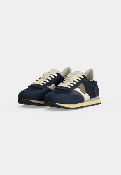 Paire de baskets bleu marine et beige à lacets avec semelles couleur crème et semelles extérieures noires texturées, positionnées côte à côte sur un fond uni.