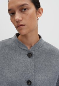 Veste en mélange de laine grise avec un col, présentant deux boutons foncés et une finition texturée. Gros plan sur la partie supérieure du vêtement.