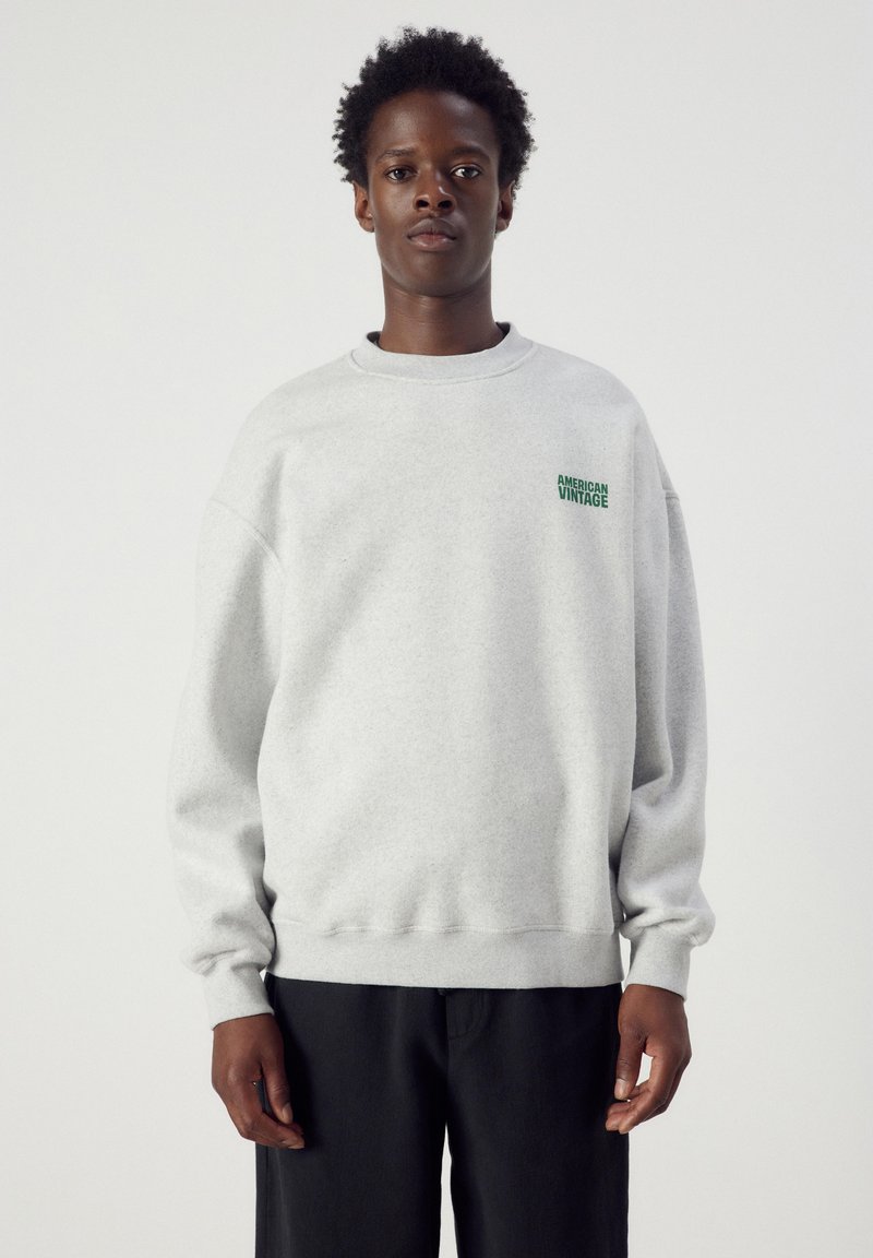Jeune homme portant un sweat-shirt gris clair avec texte vert "American Vintage" sur la poitrine, associé à un pantalon noir, debout sur fond blanc.