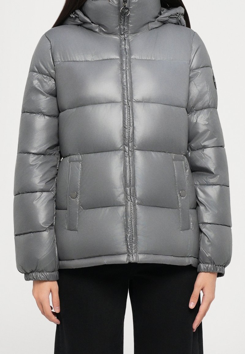 Veste d'hiver matelassée grise avec fermeture éclair avant, poches à bouton-pression, poignets élastiques et capuche, portée sur un pantalon noir.