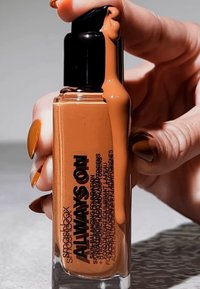 Flacon de fond de teint avec une base en verre transparent, un bouchon noir et un liquide de couleur caramel qui coule. Texte sur le côté en noir.