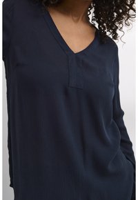 Femme portant un haut texturé bleu foncé à manches longues avec un col en V et une courte patte de boutonnage, cheveux bouclés partiellement visibles.