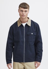 Marinblå bomulljacka med beige krage, dragkedjestängning och framficka. Har en slät textur och en figursydd design.