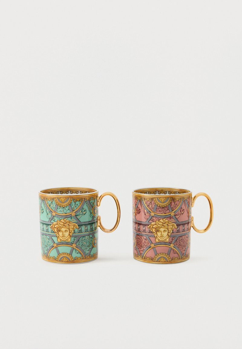 Deux tasses en céramique ornées avec des anses dorées, l'une turquoise et l'autre rose, présentant des motifs complexes en or et noir avec une tête de Méduse et des motifs géométriques.