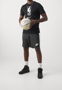 Camiseta negra de la NBA de Nike con logo, pantalones cortos grises con panel blanco, sosteniendo un balón de baloncesto beige de Nike; zapatillas altas negras, calcetines blancos.