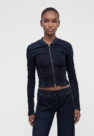 Donna che indossa una giacca aderente in denim scuro a maniche lunghe con cerniera frontale e pantaloni coordinati in denim scuro con coulisse su sfondo neutro.