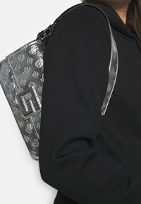 Metallisch silberne Tasche mit eingeprägten Logomustern und einem rechteckigen Hardware-Akzent. Über der Schulter getragen vor einem schwarzen Textil-Hintergrund.