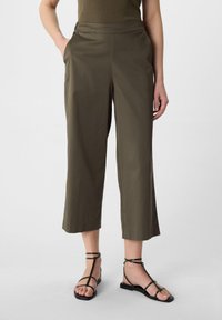 Oliwkowe spodnie 3/4 o gładkiej fakturze, z bocznymi kieszeniami i płaskim paskiem, zestawione z czarnymi sandałami na ramiączkach.