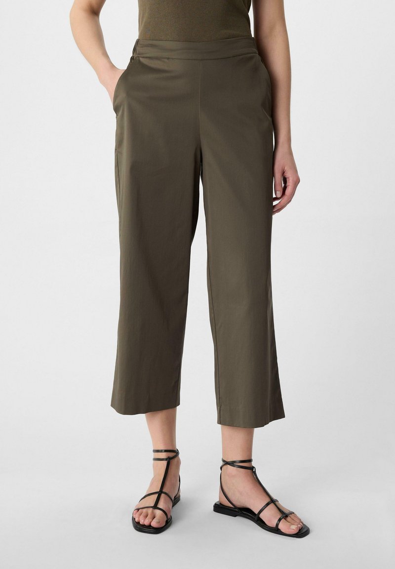 Oliwkowe spodnie 3/4 o gładkiej fakturze, z bocznymi kieszeniami i płaskim paskiem, zestawione z czarnymi sandałami na ramiączkach.