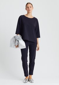 Navy oversized Oberteil mit rundem Ausschnitt, kombiniert mit eng anliegenden navyfarbenen Hosen. Hält eine silberne metallische Tasche und trägt glänzende silberne Slipper.