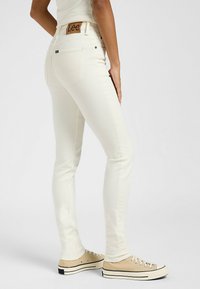 Jeans blancos ajustados de mezcla de algodón, con un parche de cuero marrón con el logo y una costura sutil, combinados con zapatillas de lona beige.