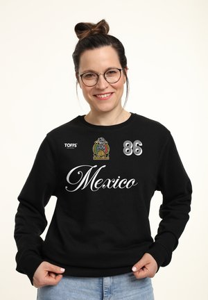Kobieta w okularach, z włosami związanymi w kok, uśmiechająca się, ubrana w czarną bluzę Mexico z logo i numerem 86, trzymająca brzeg bluzki obiema rękami.