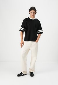 Sort kortærmet t-shirt med hvide striber på ærmerne, kombineret med lys beige bukser og sorte sneakers. Modellen står foran en hvid baggrund.