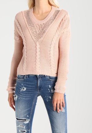 Pull en tricot rose clair avec un motif texturé, doté d'un col large et de poignets côtelés. Porté avec un jean bleu déchiré.