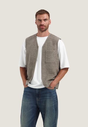 Man die een grijze vest met ritssluiting draagt over een wit t-shirt en blauwe spijkerbroek, staand met handen in de zakken tegen een effen achtergrond.