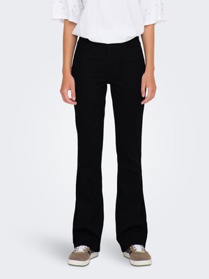 Persona che indossa pantaloni a zampa neri, top bianco a maniche corte e sneakers marroni, in piedi davanti a uno sfondo chiaro e semplice.