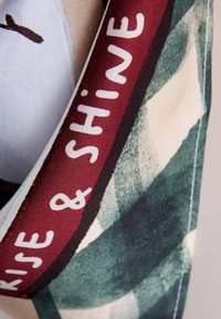 Bavlnená látka so zeleno-bielym abstraktným vzorom. Bordová stuha nesie nápis "RISE & SHINE" v bielom texte, s hladkou textúrou.