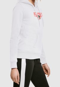 Biała bluza z kapturem i kieszenią z przodu, z napisem "PEACHES" w koralowym kolorze oraz grafiką brzoskwini. Zestawiona z czarnymi spodniami z białymi paskami.