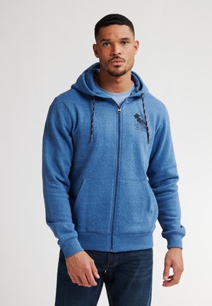 Hoodie zippé bleu avec une texture douce, cordons de serrage et poches devant. Présente un logo sur la poitrine et des poignets côtelés avec une coupe droite.