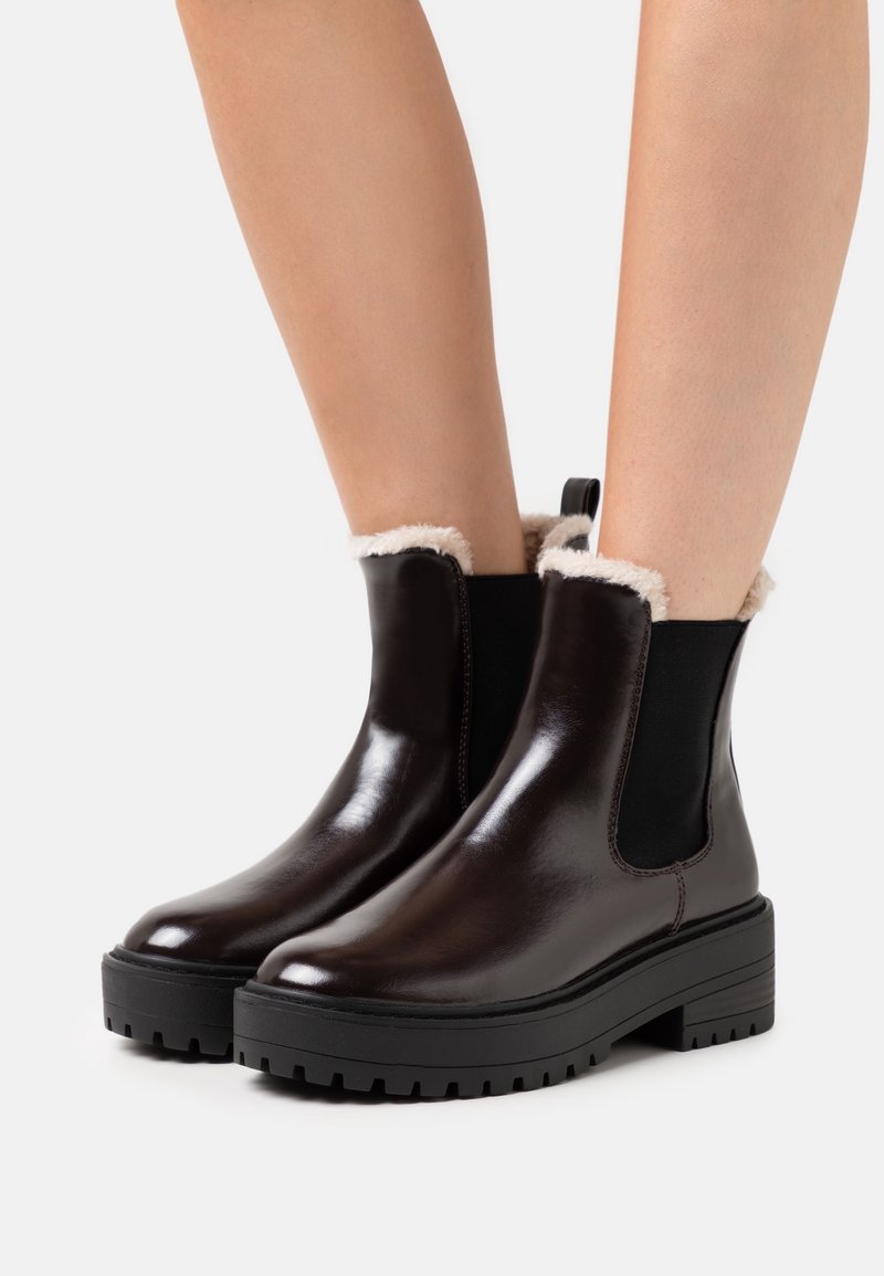 Kiillotetut tummanruskeat nilkkasandaalit, joissa on mustat elastiset sivut, tukevat mustat pohjat ja beige turkistarvikkeiden vuori, joita pidetään paljain jaloin valkoista taustaa vasten.