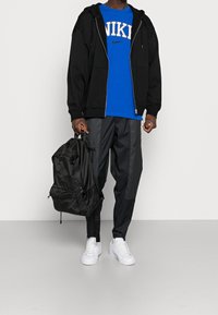 Svart zip-up hoodie över en blå Nike t-shirt, svarta tapered byxor med dragkedjor och vita sneakers. Bär en svart ryggsäck.