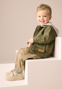 Oliivinvihreä takki harmaalla hupulla, beiget housut ja vaaleanbeiget tarrakengät. Takissa on tasku ja housuissa kuminauhakujaan.