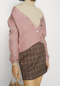 Virkat rosa cardigan med överdimensionerad krage och knappar, kombinerad med en brun rutig kjol. Beige polotröja under, håller en beige handväska.