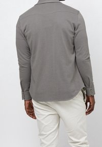 Harmaa pitkähihainen paita, jossa on kaulus, nappisalaiset hihat ja pyöristetty helma. Sujuva, pehmeä kangas, jossa on minimaalista yksityiskohtaisuutta takana.