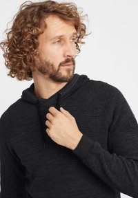 Hoodie aus strukturiertem schwarzem Stoff mit gerippten Bündchen, ausgestattet mit einer Kordelzugkapuze und einem runden Logopatch auf der linken Schulter.