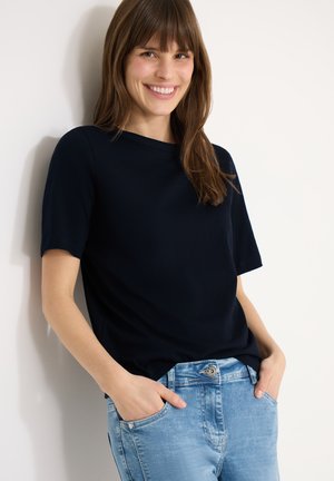 Smilende kvinde med glat brunt hår iført sort kortærmet skjorte og lyseblå jeans, står med hænderne i lommerne mod en hvid væg.