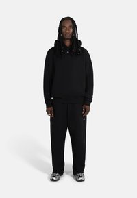 Svart hettegenser med løs passform, kombinert med matchende svarte joggebukser. Har minimal merkevareprofil og grå sneakers med detaljer.