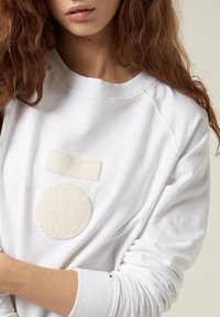 Donna che indossa un felpa bianca con patch testurizzati a forma di cerchio e rettangolo bianchi sul petto, con lunghi capelli castani mossi visibili.