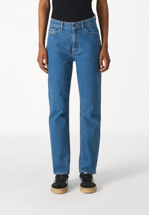 Person iført blå jeans med lige ben og sorte sneakers med hvide striber, stående mod en hvid baggrund.