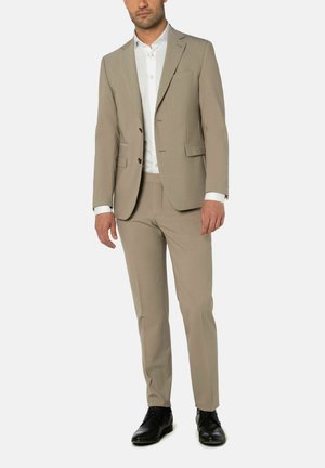 Homme portant un costume beige à deux boutons avec un pantalon assorti, une chemise blanche et des chaussures noires, debout devant un fond uni.