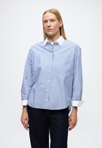 Camicia a bottoni con righe verticali blu e bianche, colletto bianco e dettagli sui polsini. Presenta una tasca applicata sul lato sinistro.