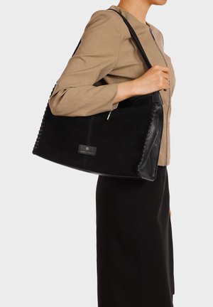 Donna con giacca beige e gonna nera che tiene una grande borsa nera in pelle sulla spalla su sfondo bianco.