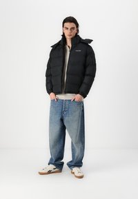 Μαύρο μπουφάν με φερμουάρ, με κουκούλα και ελαστικούς μανσέτες. Φορεμένο πάνω από ένα ανοιχτόχρωμο πουλόβερ και συνδυασμένο με χαλαρές μπλε τζιν και αθλητικά παπούτσια.