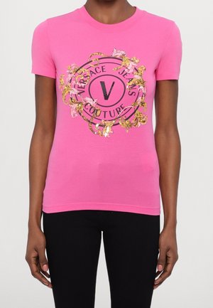 Rosa T-Shirt mit kurzen Ärmeln, verziert mit einem kreisförmigen Logo in Schwarz und Gold mit floralen Akzenten und Glitzerdetails in der Mitte.