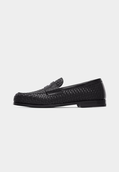 Mocassin en cuir tressé noir avec un petit talon et bout mocassin, présentant une sangle horizontale sur le dessus sur un fond blanc uni.