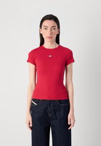 Femme portant un t-shirt rouge ajusté à manches courtes avec un petit logo, associé à un jean large bleu foncé, se tenant devant un fond uni.